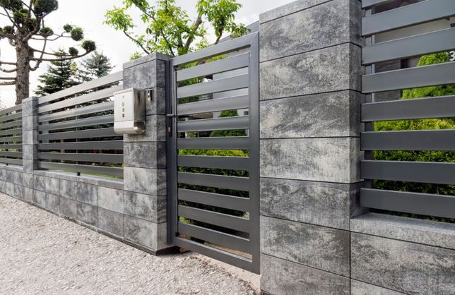 Nowoczesna brama wjazdowa i przęsła ogrodzeniowe z poziomymi, metalowymi profilami, osadzone na słupkach i podmurówce z szarego, imitującego kamień betonu architektonicznego, z widoczną skrzynką...