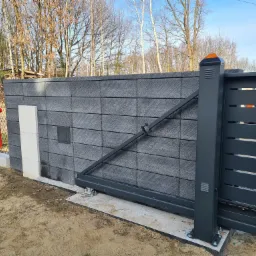 Nowoczesna, przesuwna brama wjazdowa z szarymi, poziomymi panelami i słupkiem z pomarańczowym światłem, połączona z murem z szarej kostki łupanej, widoczny hydrant i siatka ogrodzeniowa w tle.