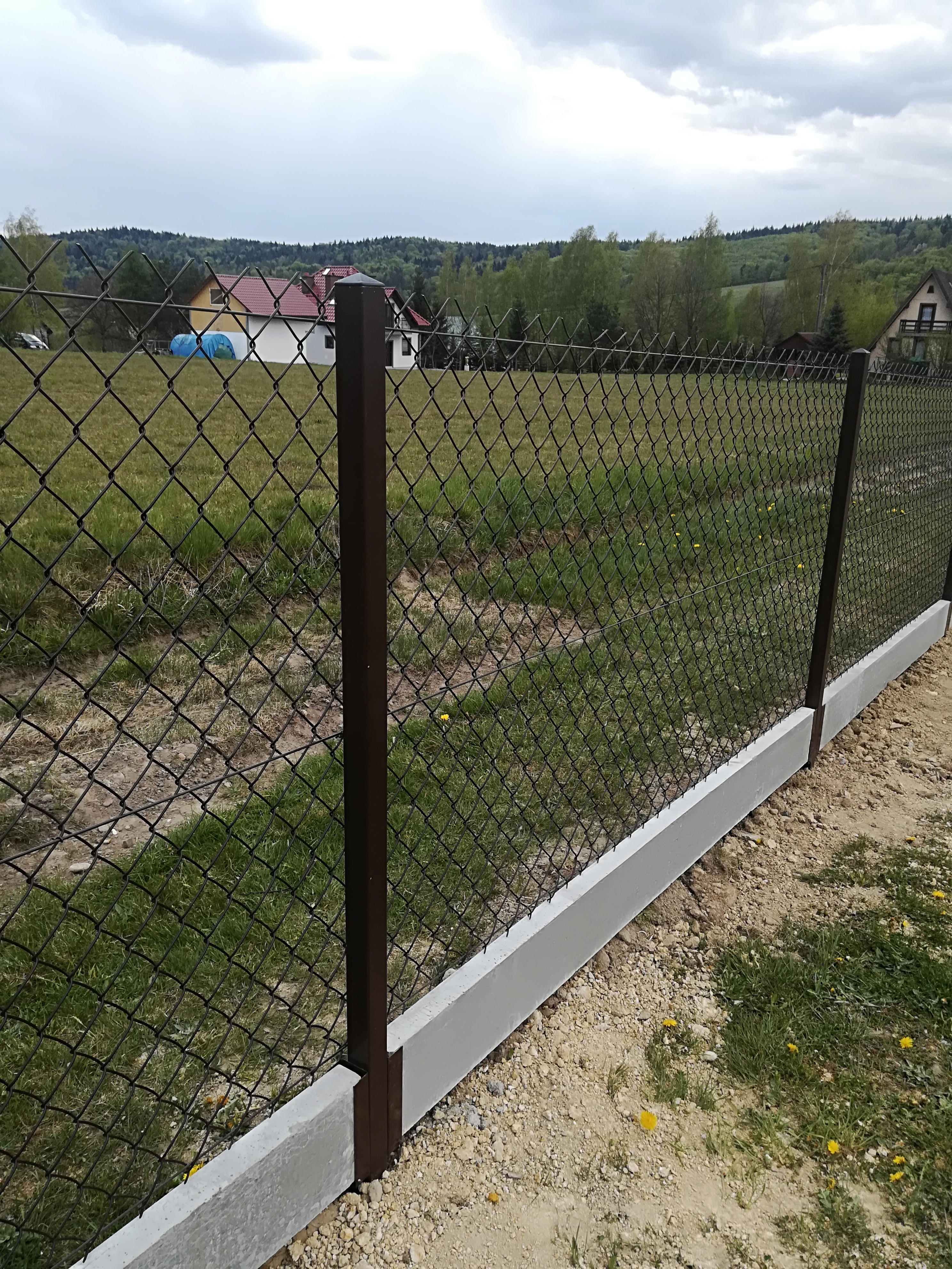 Nowo postawione ogrodzenie z siatki z brązowymi słupkami i betonową podmurówką na tle zielonego krajobrazu z domami.