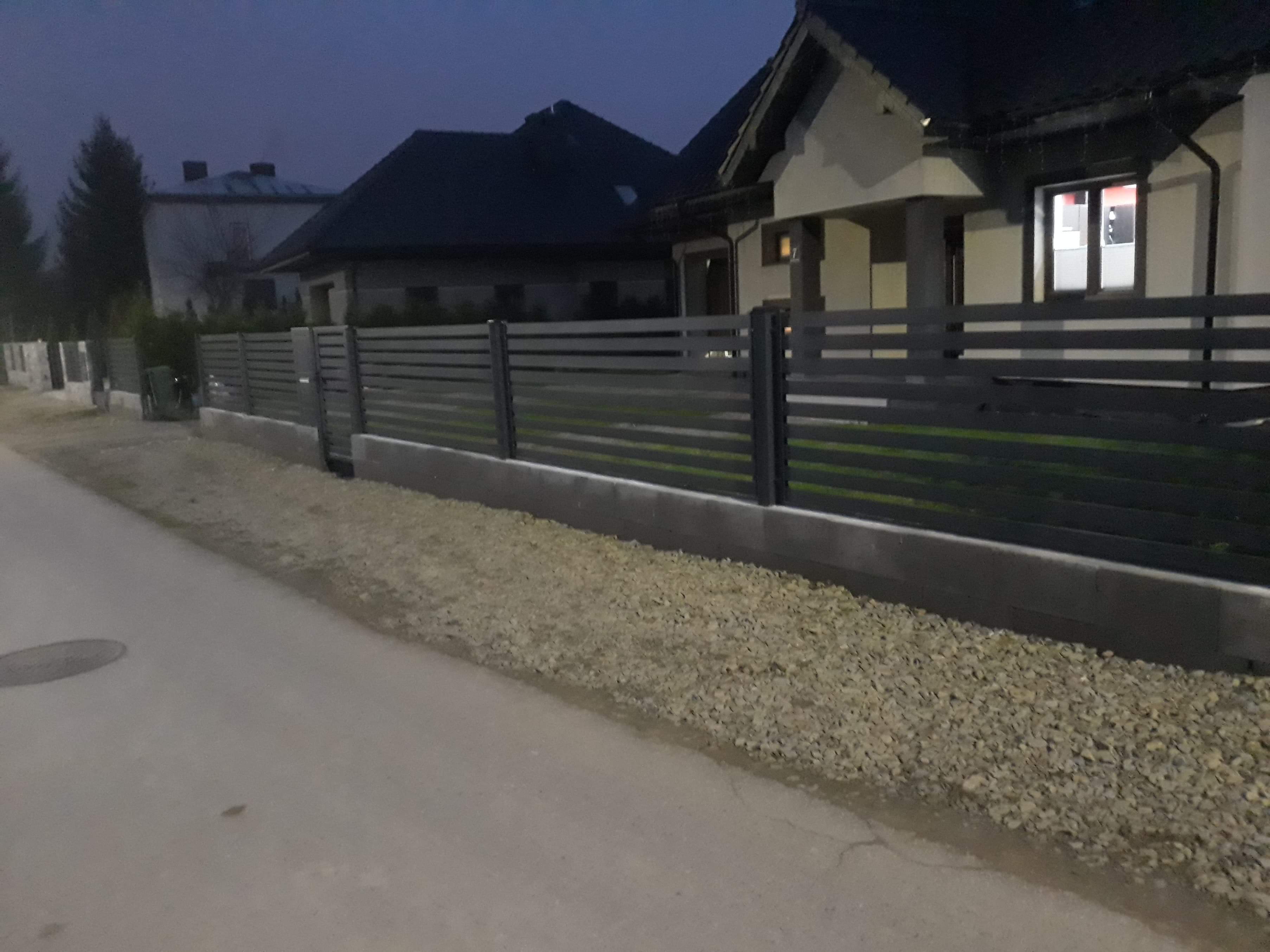 Nowoczesne, poziome, szare ogrodzenie z paneli metalowych zamontowane na betonowej podmurówce przed domem jednorodzinnym oświetlonym wieczornym światłem.