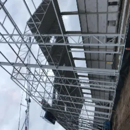 Wznoszenie konstrukcji stalowej hali z widocznym szkieletem dachu i ścian, z dźwigiem w tle i robotnikami na wysokości.