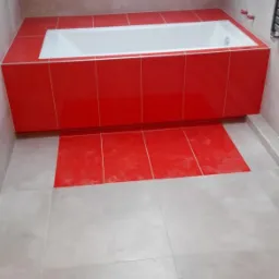 Wykończona łazienka z wanną obudowaną jaskrawo czerwoną glazurą, dopasowaną do fragmentu podłogi przy wannie. Reszta podłogi wyłożona jest jasnymi, kwadratowymi płytkami. Na ścianach szara glazura.