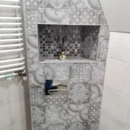 Szare płytki w łazience z dekoracyjnym wzorem, wnęka z mozaiką i złotym dyfuzorem zapachowym, chromowany przycisk spłuczki, biały grzejnik drabinkowy po lewej stronie.