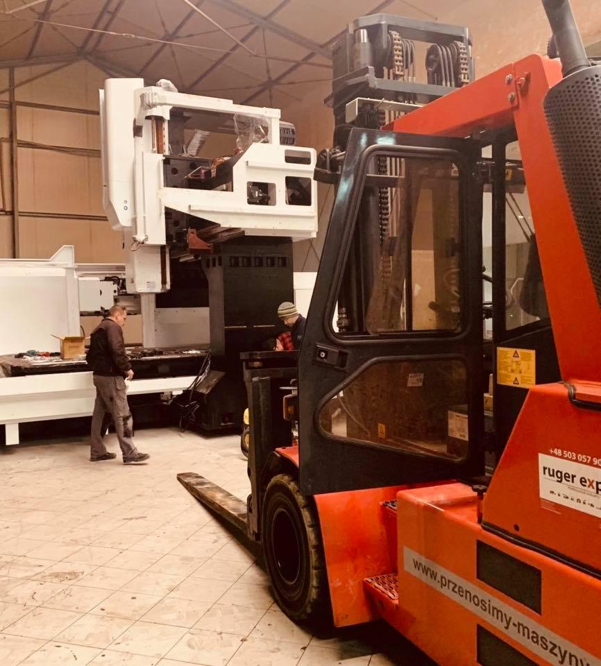 Pomarańczowy wózek widłowy w hali produkcyjnej, transportujący dużą białą maszynę CNC; widoczni dwaj mężczyźni nadzorujący operację.