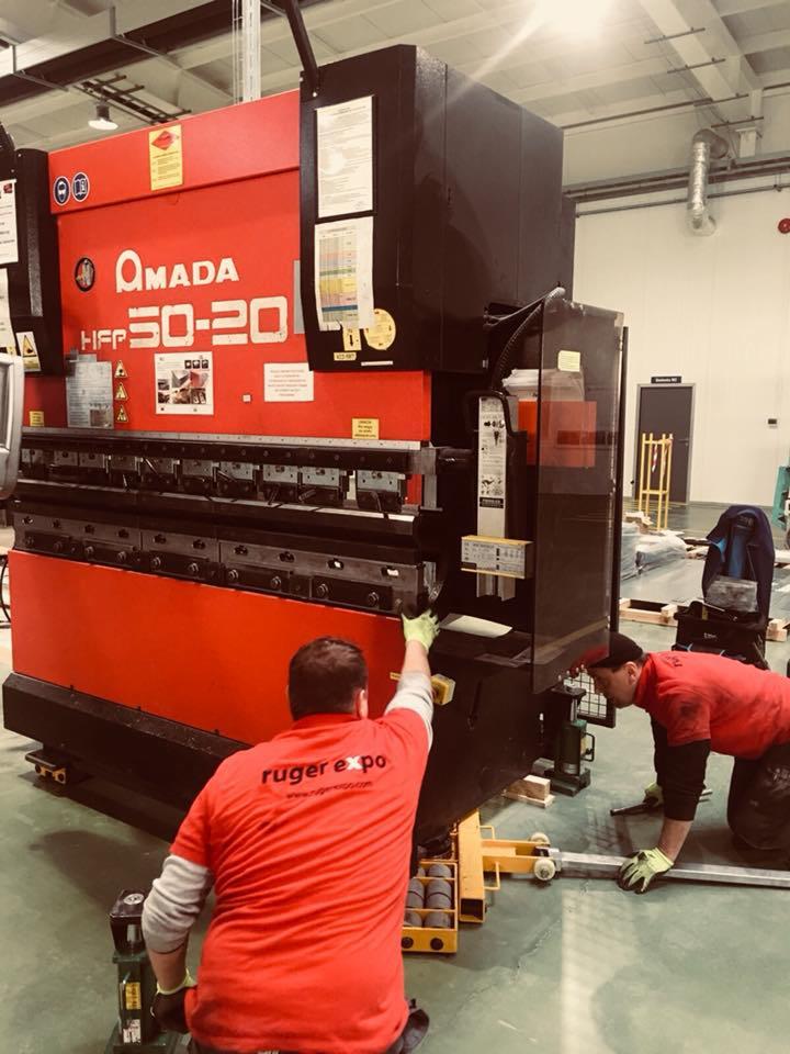 Dwóch mężczyzn w czerwonych koszulkach z logo 'ruger expo' instaluje dużą, czerwoną maszynę Amada HFP 50-20 w hali przemysłowej.