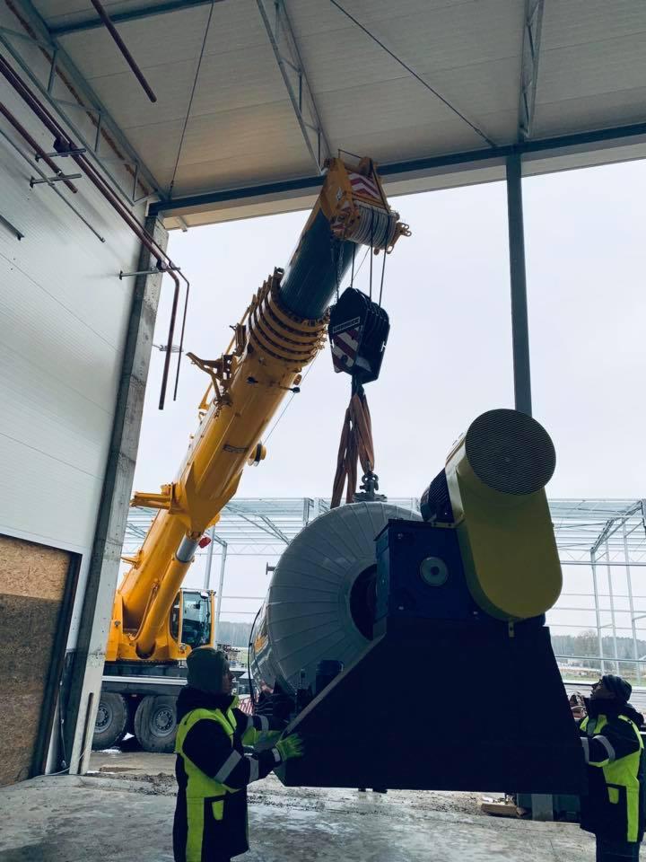 Żółty dźwig podnosi szaro-czarny, cylindryczny element wewnątrz hali z konstrukcją stalową, pracownicy w kamizelkach odblaskowych nadzorują operację.