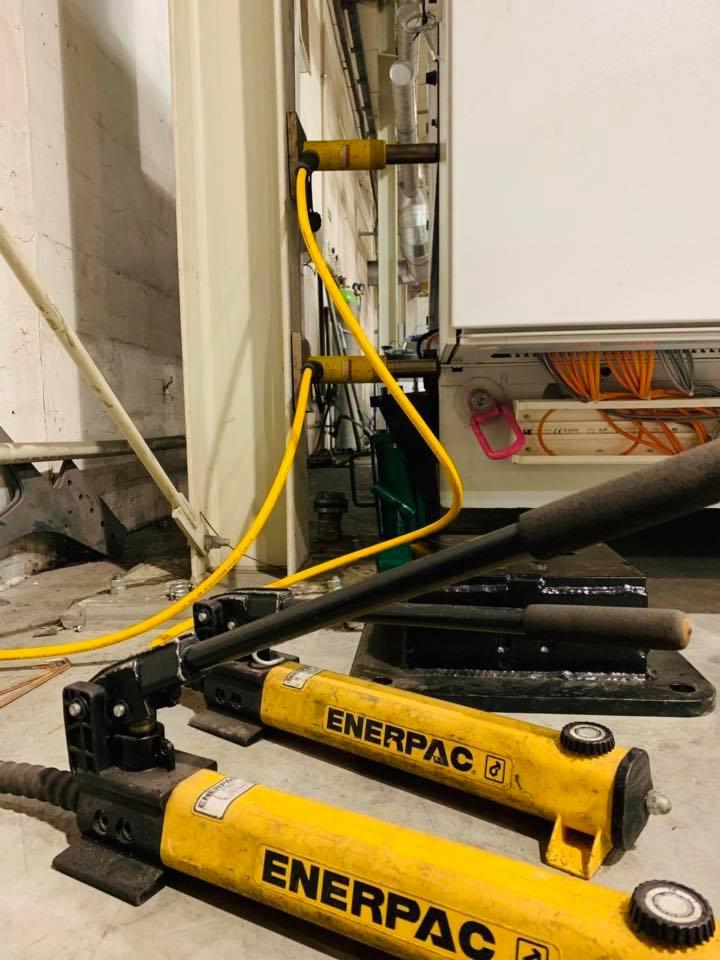 Dwa żółte siłowniki hydrauliczne marki Enerpac leżące na podłodze, z jednym podłączonym do kolumny i części maszyny w tle, z widocznymi przewodami hydraulicznymi.