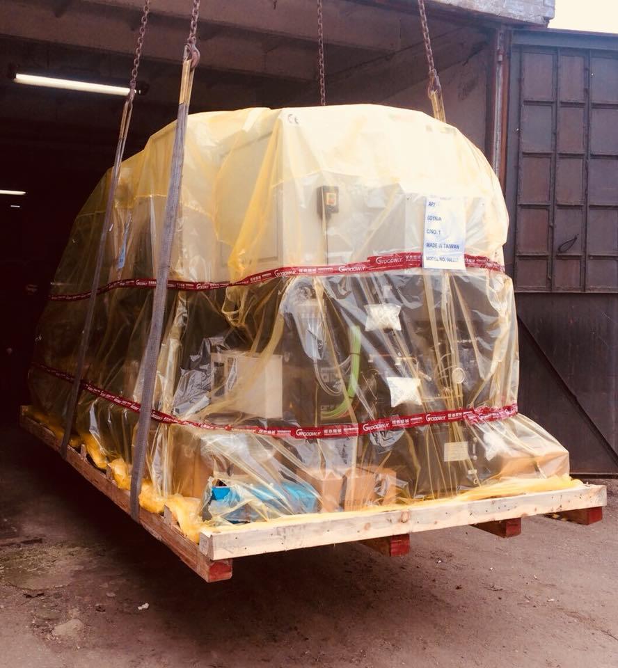 Maszyna przemysłowa zapakowana w folię ochronną, umieszczona na drewnianej palecie, podwieszona na łańcuchach w magazynie, widoczne oznaczenia 'Made in Taiwan'.