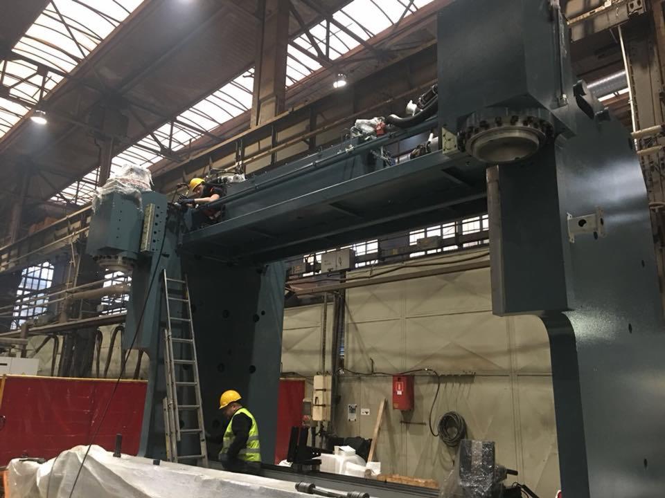 Dwóch pracowników w kaskach i kamizelkach odblaskowych pracuje nad montażem ogromnej, szarej obrabiarki przemysłowej w hali fabrycznej, jeden na drabinie, drugi na górnej platformie maszyny.