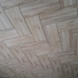 Układanie podłogi z płytek imitujących drewno w jodełkę, widoczny fragment surowego sufitu i ściany w trakcie prac wykończeniowych.