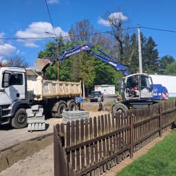 Bojarczuk Mariusz -Koparki - Koparka Atlas ładuje ziemię do wywrotki MAN na placu budowy, w tle widoczni pracownicy i sterta kostki brukowej, a w pierwszym planie drewniany płot.