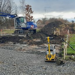 Bojarczuk Mariusz -Koparki - Niebieska koparka Atlas wykopuje ziemię na działce, obok ubijarka wibracyjna, niwelator laserowy na statywie i samochód dostawczy, wstępne utwardzanie drogi tłuczniem.