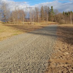 Bojarczuk Mariusz -Koparki - Świeżo utwardzona żwirowa droga gruntowa prowadząca w kierunku lasu, widoczne ślady opon po prawej stronie, błękitne niebo z chmurami w tle.