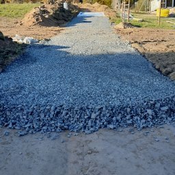 Bojarczuk Mariusz -Koparki - Przygotowanie podłoża: warstwa szarego kruszywa na utwardzonej drodze gruntowej, widok w perspektywie z nowo budowanym domem w tle.