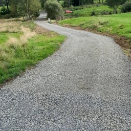 Bojarczuk Mariusz -Koparki - Wąska, szutrowa droga wijąca się przez zielone łąki i lasy, z czerwonym samochodem w tle i błękitnym niebem z chmurami.