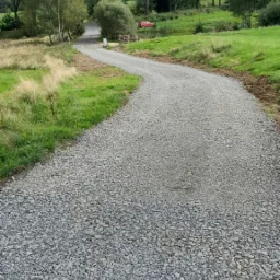 Wąska, szutrowa droga wijąca się przez zielone łąki i lasy, z czerwonym samochodem w tle i błękitnym niebem z chmurami.