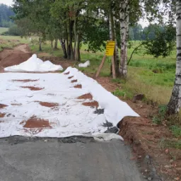 Budowa drogi gruntowej: warstwa geowłókniny na podsypce, widoczny wykop i tablica ostrzegawcza w tle.