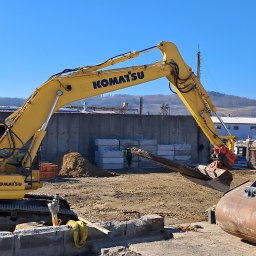 Bojarczuk Mariusz -Koparki - Żółta koparka Komatsu PC 210 Lci z hydraulicznym młotem udarowym i łyżką obok, na placu budowy z widoczną ziemią i betonowymi elementami, w tle budynki przemysłowe i wzgórza.