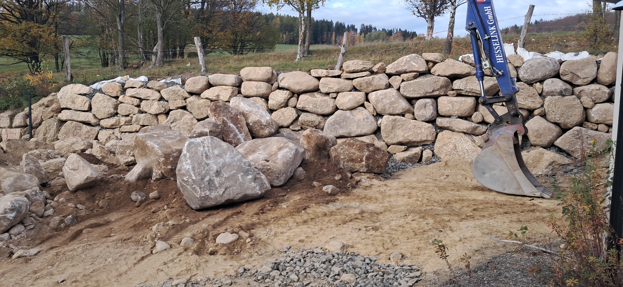 Budowa kamiennego muru oporowego z użyciem koparki Hesselink GmbH, duże kamienie na ziemi przed murem.