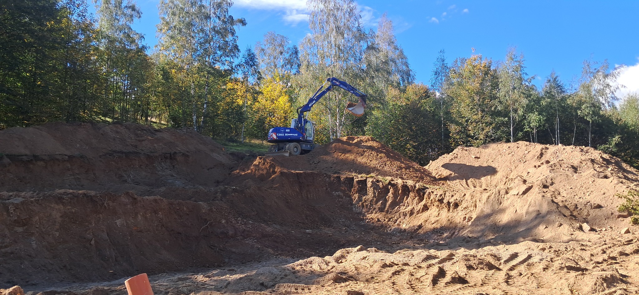 Niebieska koparka Atlas 1305 M w Jeleniej Górze, podczas prac ziemnych na tle lasu i błękitnego nieba.