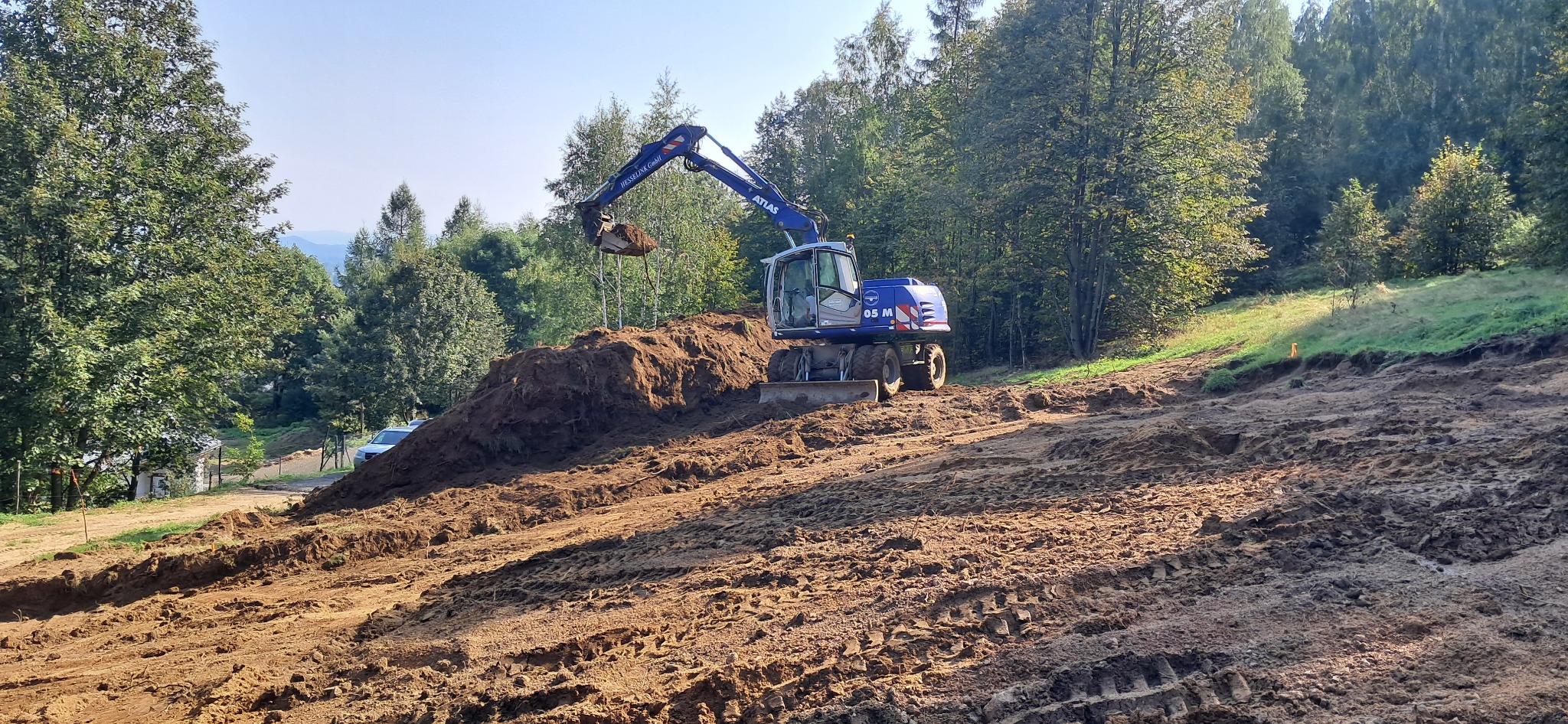 Niebieska koparka Atlas 105 M nabiera ziemię na nierównym terenie w słoneczny dzień, w tle drzewa i częściowo widoczny biały samochód.