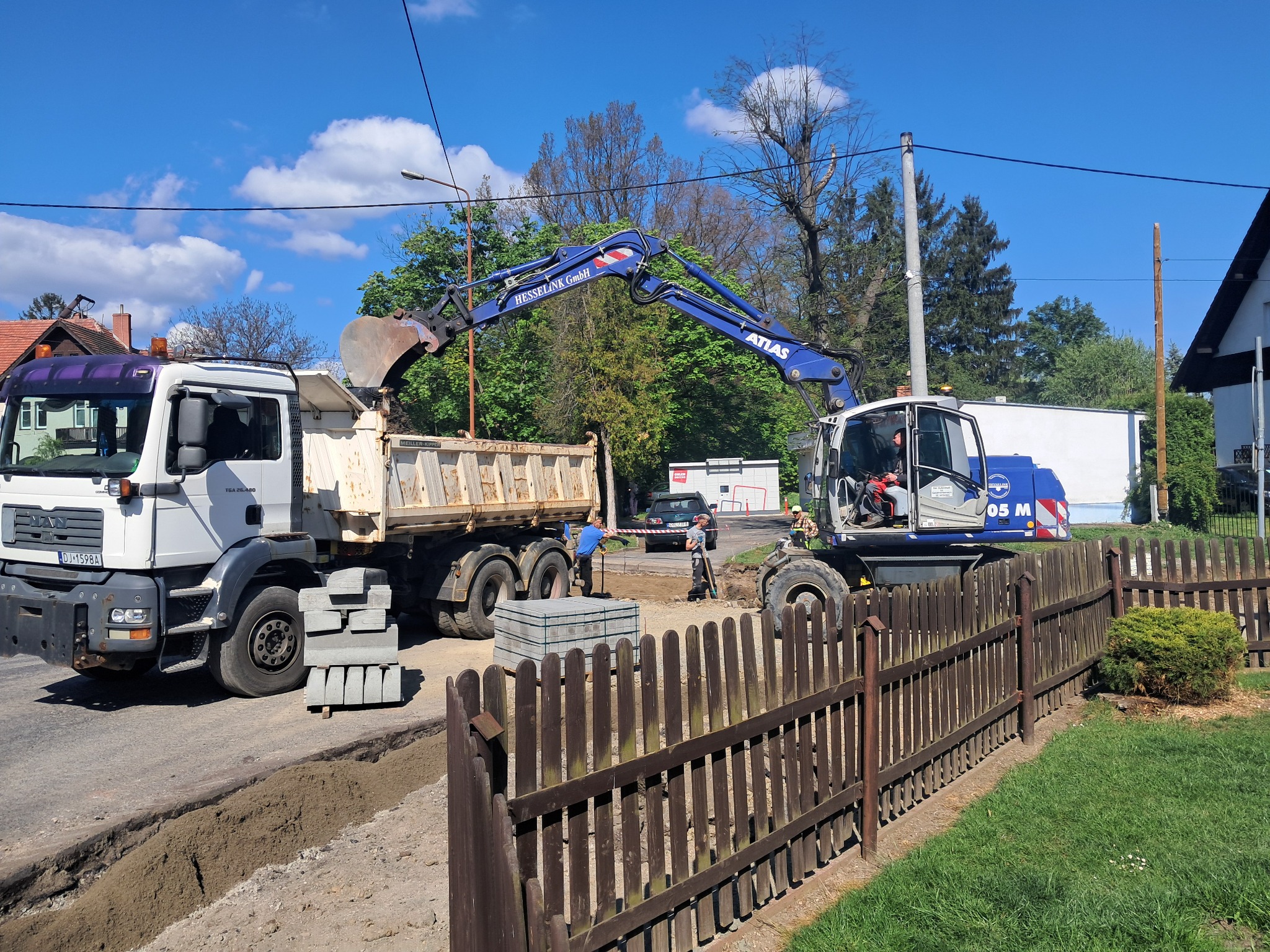 Koparka Atlas ładuje ziemię do wywrotki MAN na placu budowy, w tle widoczni pracownicy i sterta kostki brukowej, a w pierwszym planie drewniany płot.