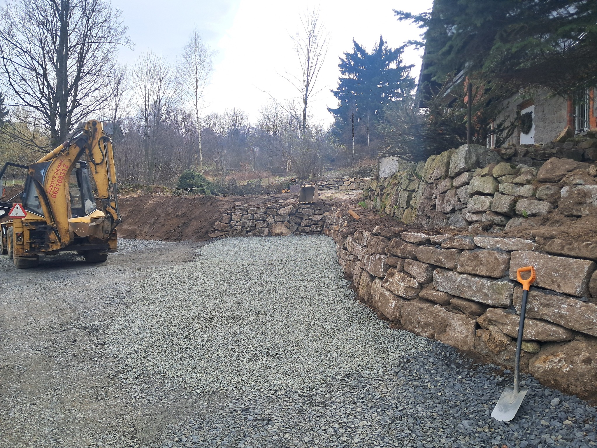 Żółta koparko-ładowarka przy nowo utworzonej kamiennej ścianie oporowej i utwardzonej powierzchni żwirowej, widoczna łopata oparta o mur.