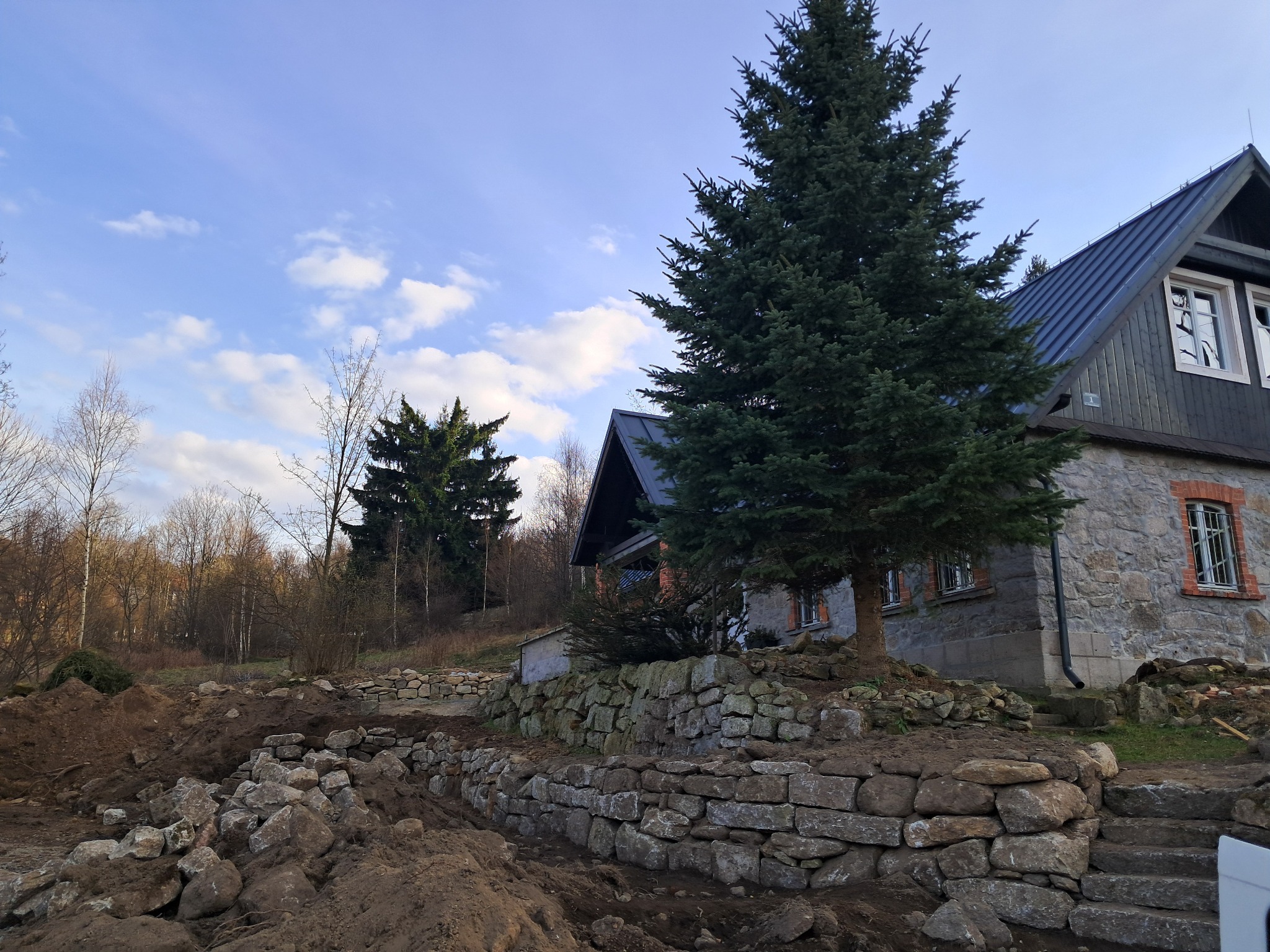 Kamienna ściana oporowa w trakcie budowy, surowy teren, dom z kamienia w tle, iglaste drzewo, błękitne niebo z chmurami.