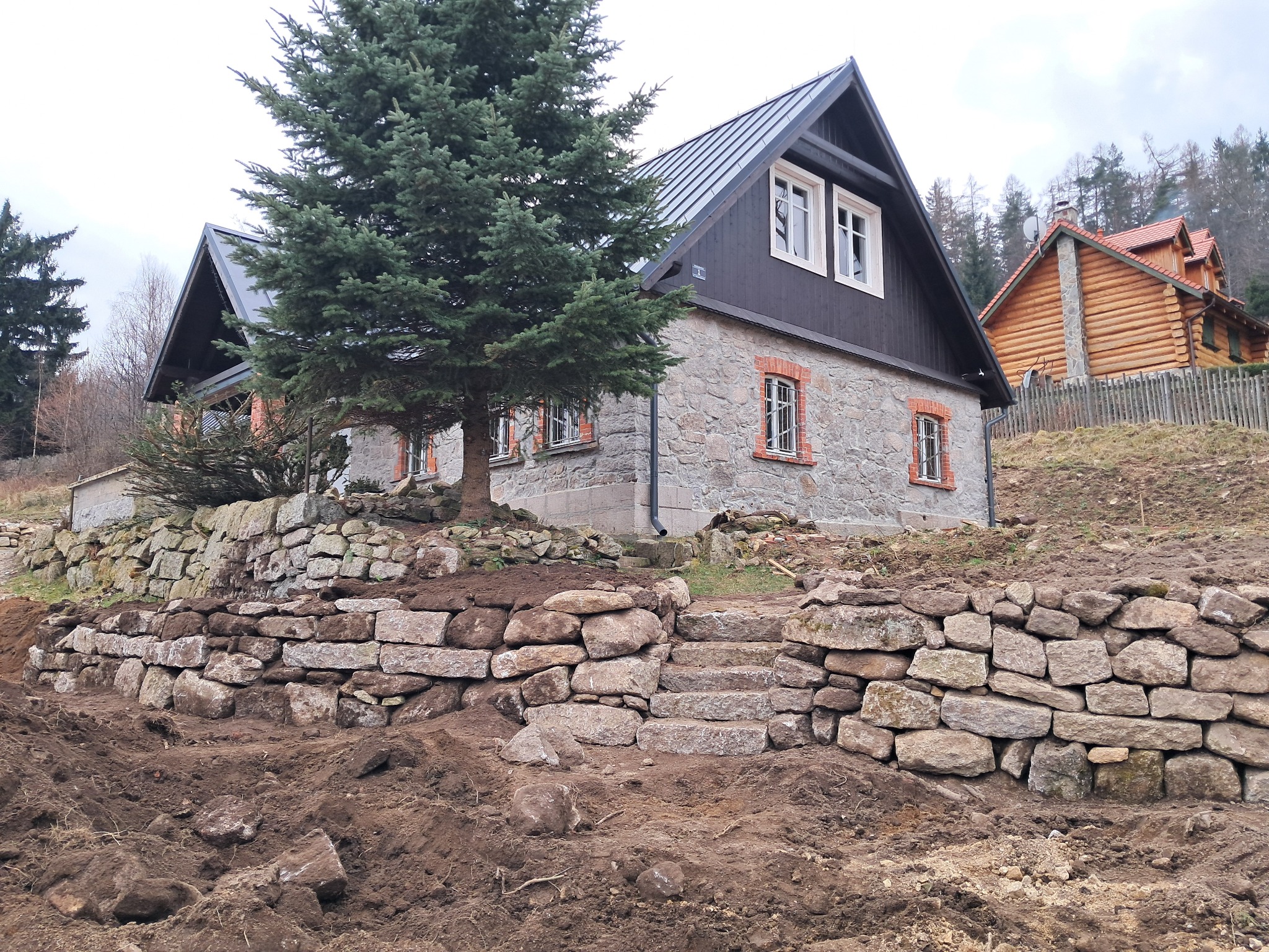 Kamienny murek oporowy zintegrowany ze schodami prowadzącymi do domu w stylu rustykalnym, z widoczną świeżo przekopaną ziemią u podstawy.