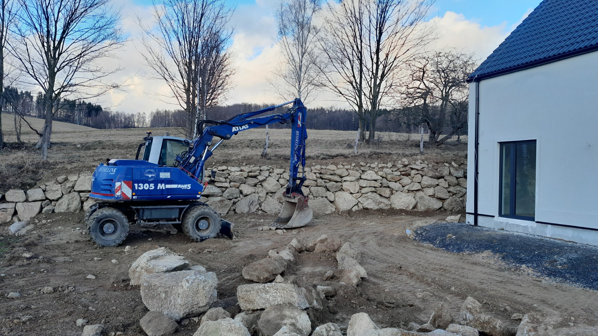 Niebieska koparka Atlas 1305 M na placu budowy, w tle kamienny mur oporowy i fragment białego budynku z ciemnym oknem, teren przygotowany pod dalsze prace.