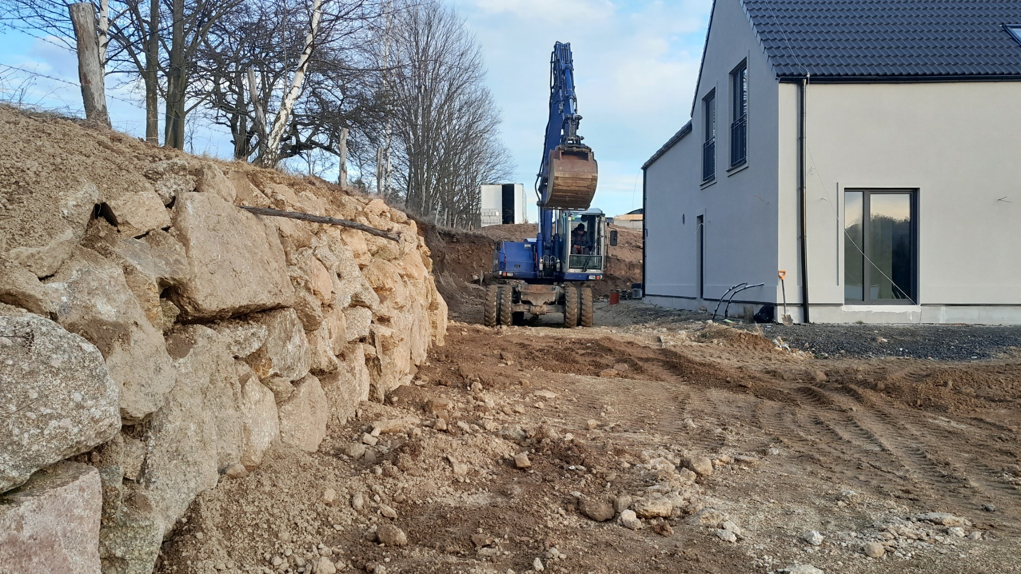 Niebieska koparka na placu budowy, widoczny wykop ziemny, kamienny mur oporowy i fragment nowoczesnego domu w tle.