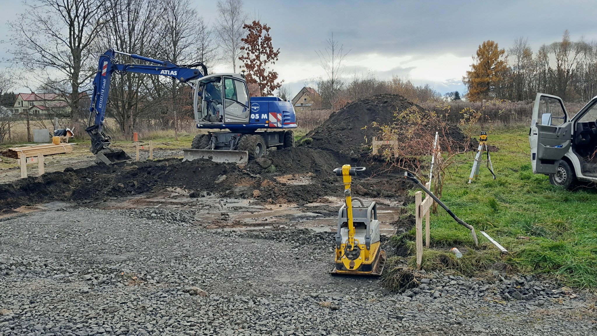 Niebieska koparka Atlas wykopuje ziemię na działce, obok ubijarka wibracyjna, niwelator laserowy na statywie i samochód dostawczy, wstępne utwardzanie drogi tłuczniem.