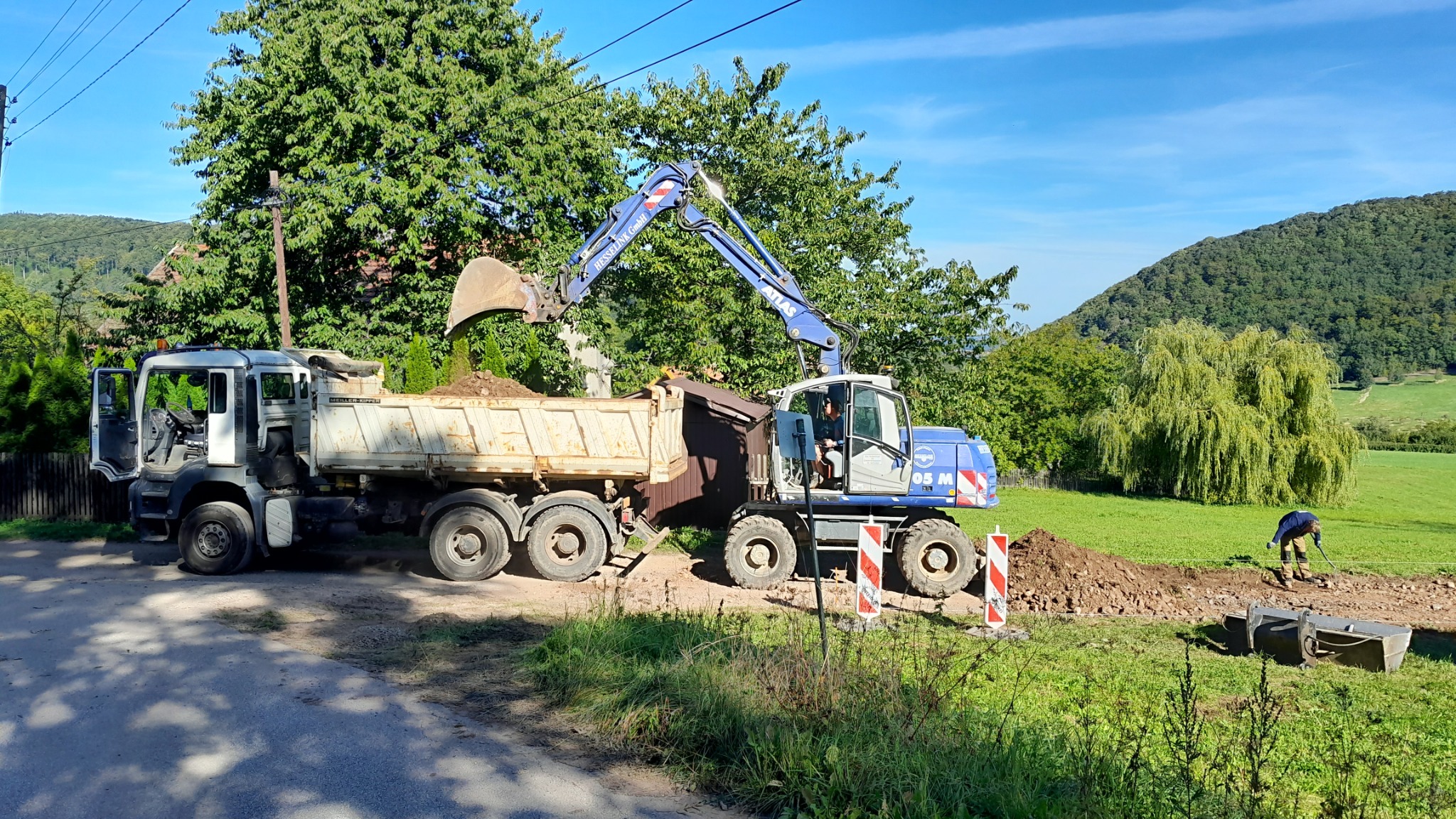 Koparka Atlas załadowuje ziemię do wywrotki na tle zielonego krajobrazu, w oddali widać pracownika z łopatą.