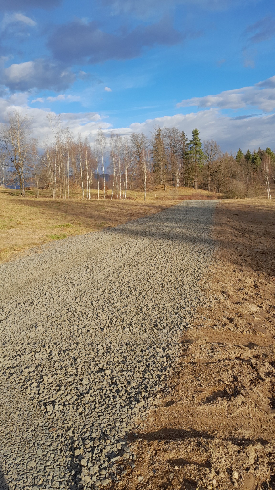 Świeżo utwardzona żwirowa droga gruntowa prowadząca w kierunku lasu, widoczne ślady opon po prawej stronie, błękitne niebo z chmurami w tle.