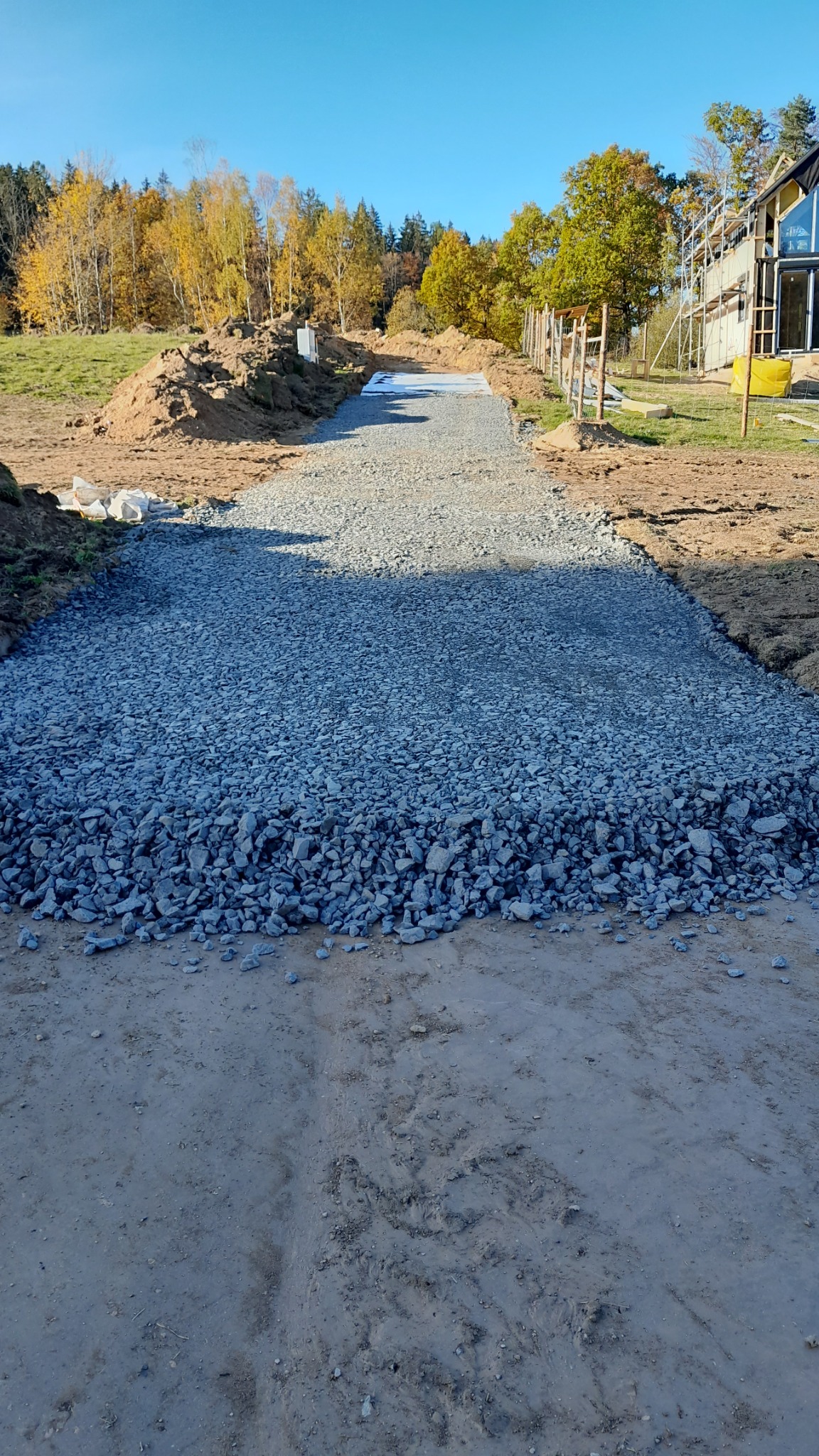 Przygotowanie podłoża: warstwa szarego kruszywa na utwardzonej drodze gruntowej, widok w perspektywie z nowo budowanym domem w tle.
