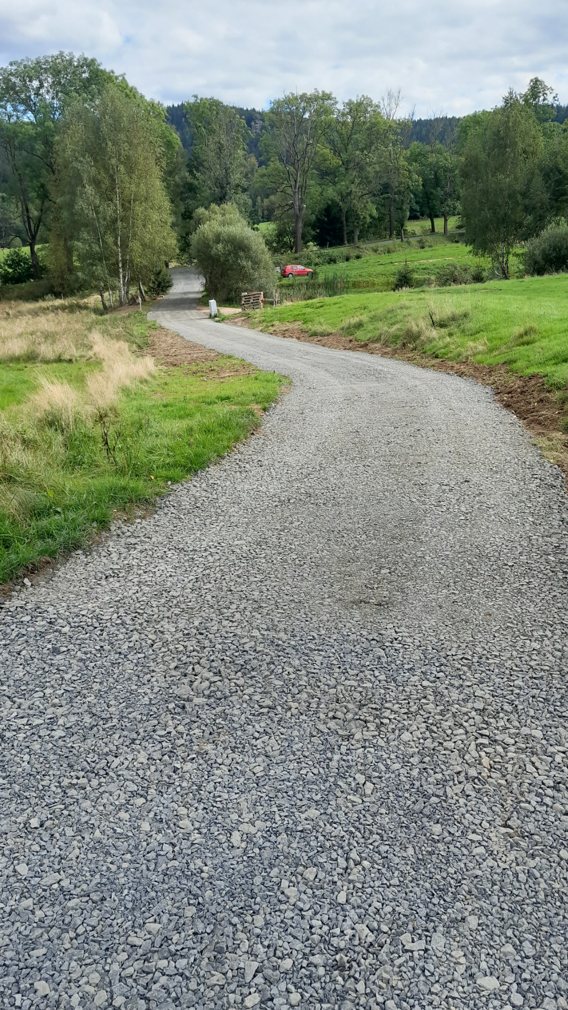 Wąska, szutrowa droga wijąca się przez zielone łąki i lasy, z czerwonym samochodem w tle i błękitnym niebem z chmurami.