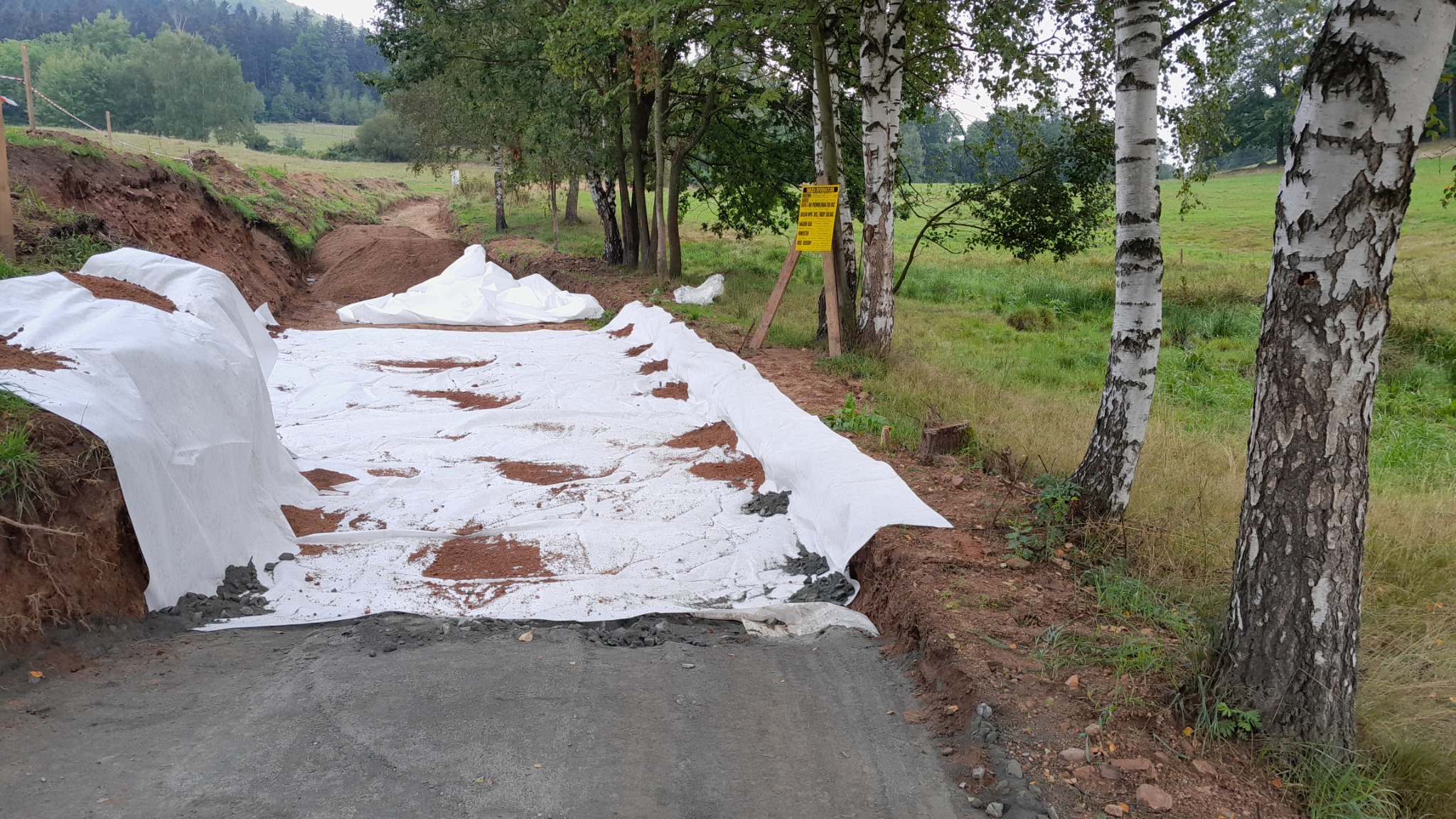 Budowa drogi gruntowej: warstwa geowłókniny na podsypce, widoczny wykop i tablica ostrzegawcza w tle.