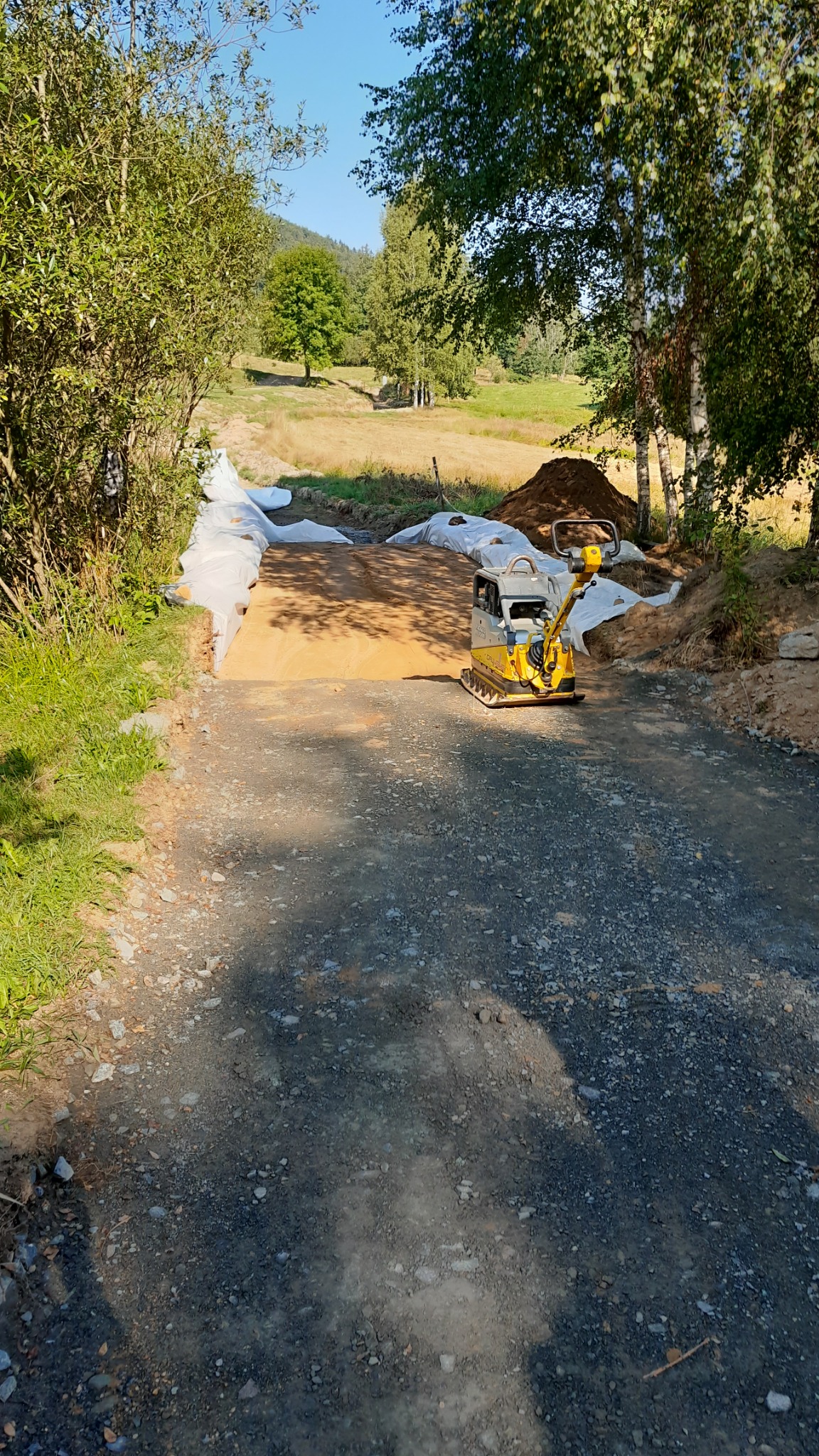 Żółta zagęszczarka obok utwardzonej powierzchni z widoczną warstwą geowłókniny i pryzmą ziemi w tle, na tle zieleni.