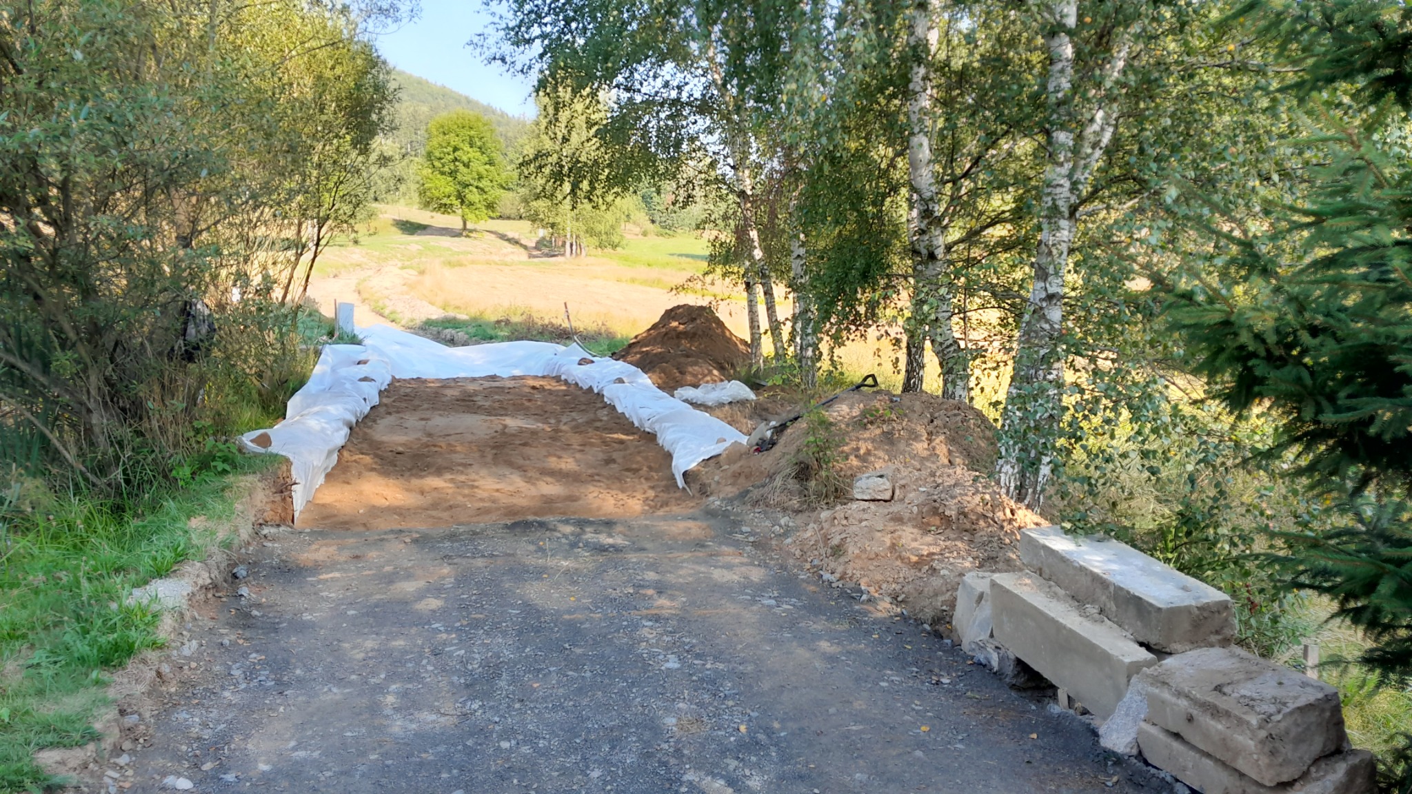 Przygotowanie podłoża drogi gruntowej, zabezpieczone folią, z widocznymi hałdami ziemi i betonowymi elementami obrzeży w malowniczym wiejskim otoczeniu.