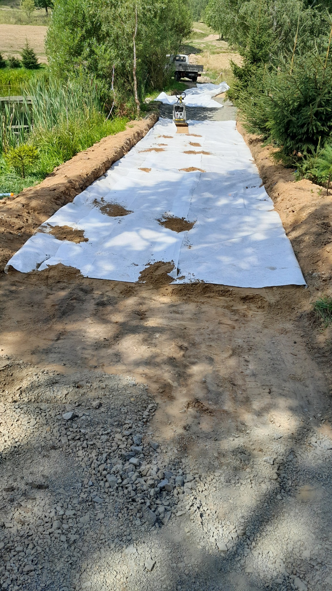 Przygotowanie podłoża pod drogę: biała geowłóknina rozłożona na ubitym żwirze, z widocznymi śladami piasku i ubijarką w tle.