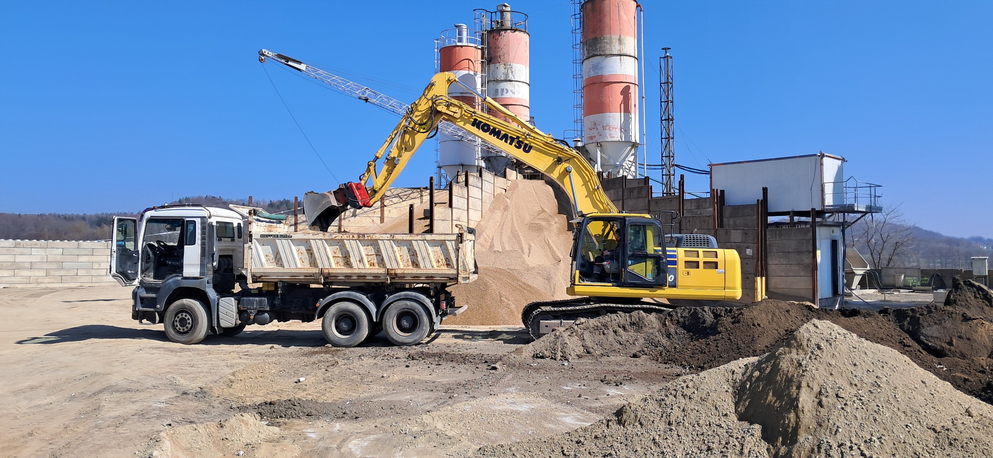 Żółta koparka Komatsu ładuje piasek do ciężarówki na tle silosów i błękitnego nieba.