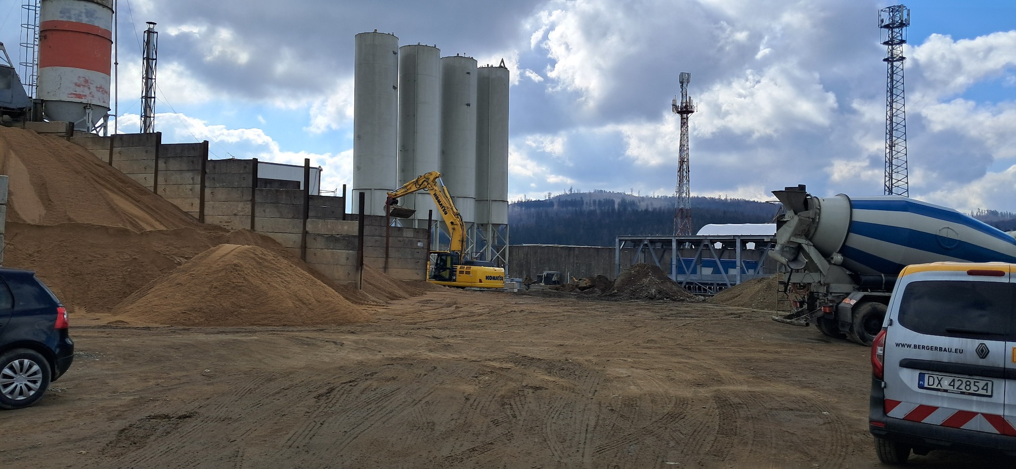 Żółta koparka Komatsu nabiera piasek na tle silosów do betonu, z boku stoi betoniarka i samochód dostawczy, w tle widoczne wzgórza i pochmurne niebo.
