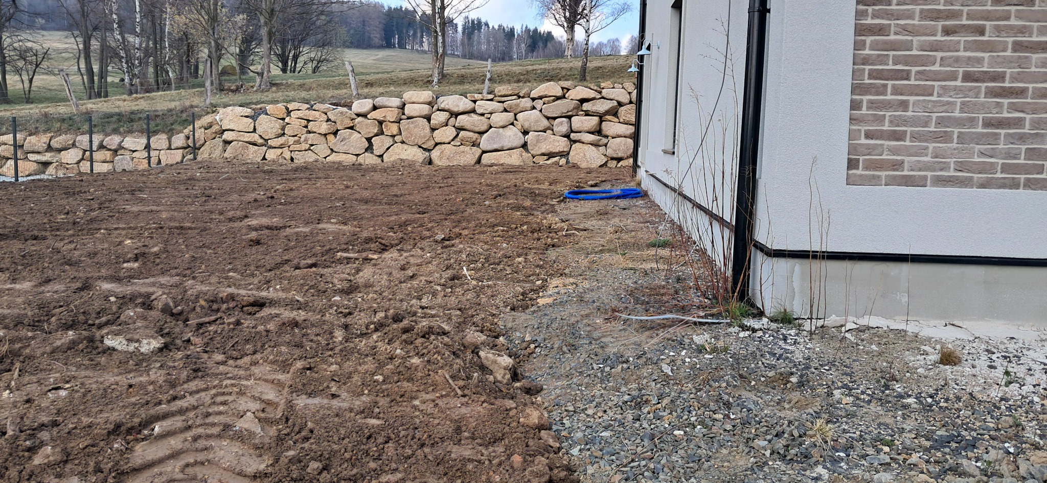 Teren przed budynkiem po pracach ziemnych, widoczne ślady opon, usypana kamienna ścieżka wzdłuż ściany budynku i kamienny murek oporowy w tle.