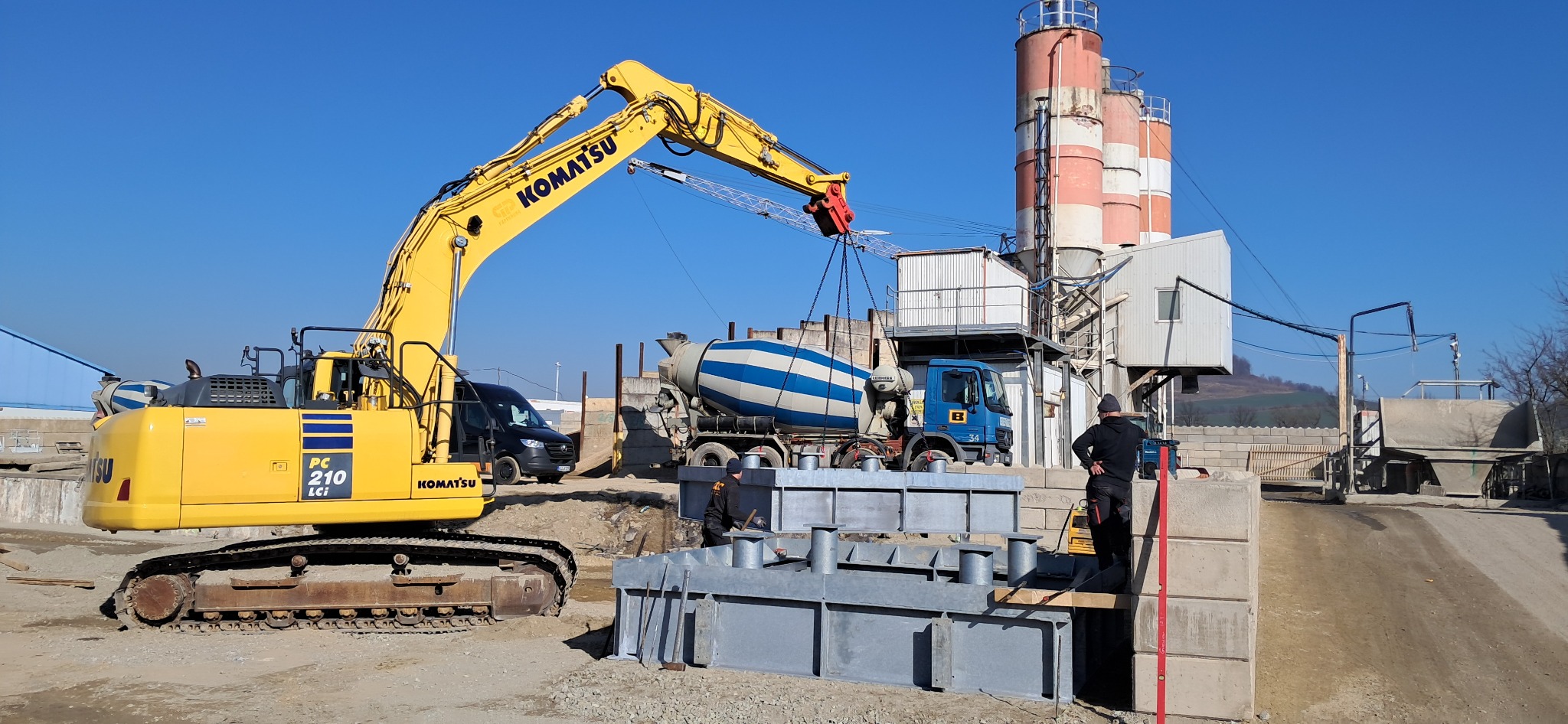 Żółta koparka Komatsu podnosi stalową konstrukcję łańcuchami, w tle betoniarka i silosy do cementu, pracownicy budowlani przy pracy.