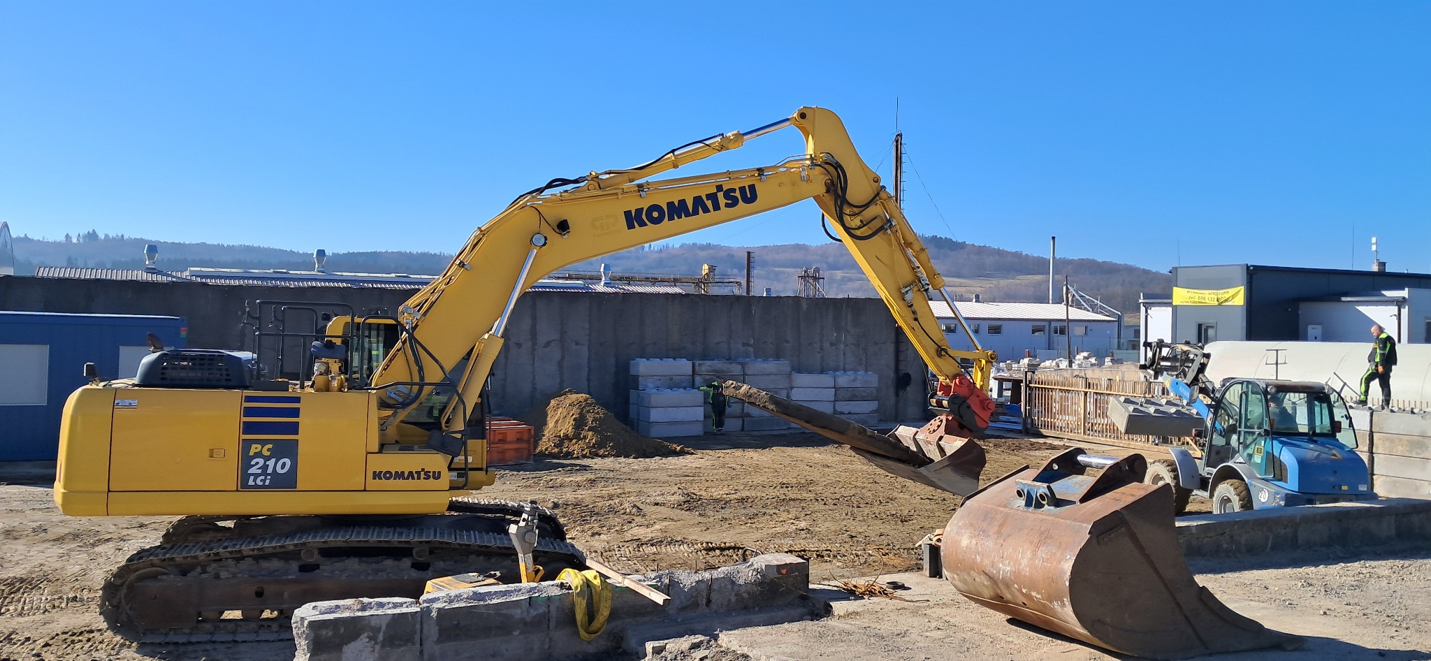 Żółta koparka Komatsu PC 210 Lci z hydraulicznym młotem udarowym i łyżką obok, na placu budowy z widoczną ziemią i betonowymi elementami, w tle budynki przemysłowe i wzgórza.