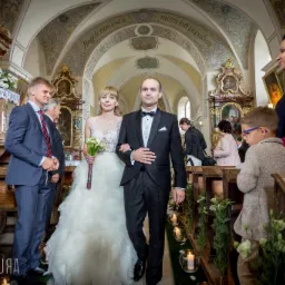 Para młoda, panna młoda w białej sukni z bukietem kalii i pan młody w czarnym smokingu, idą w kościele udekorowanym kwiatami, wśród gości weselnych.