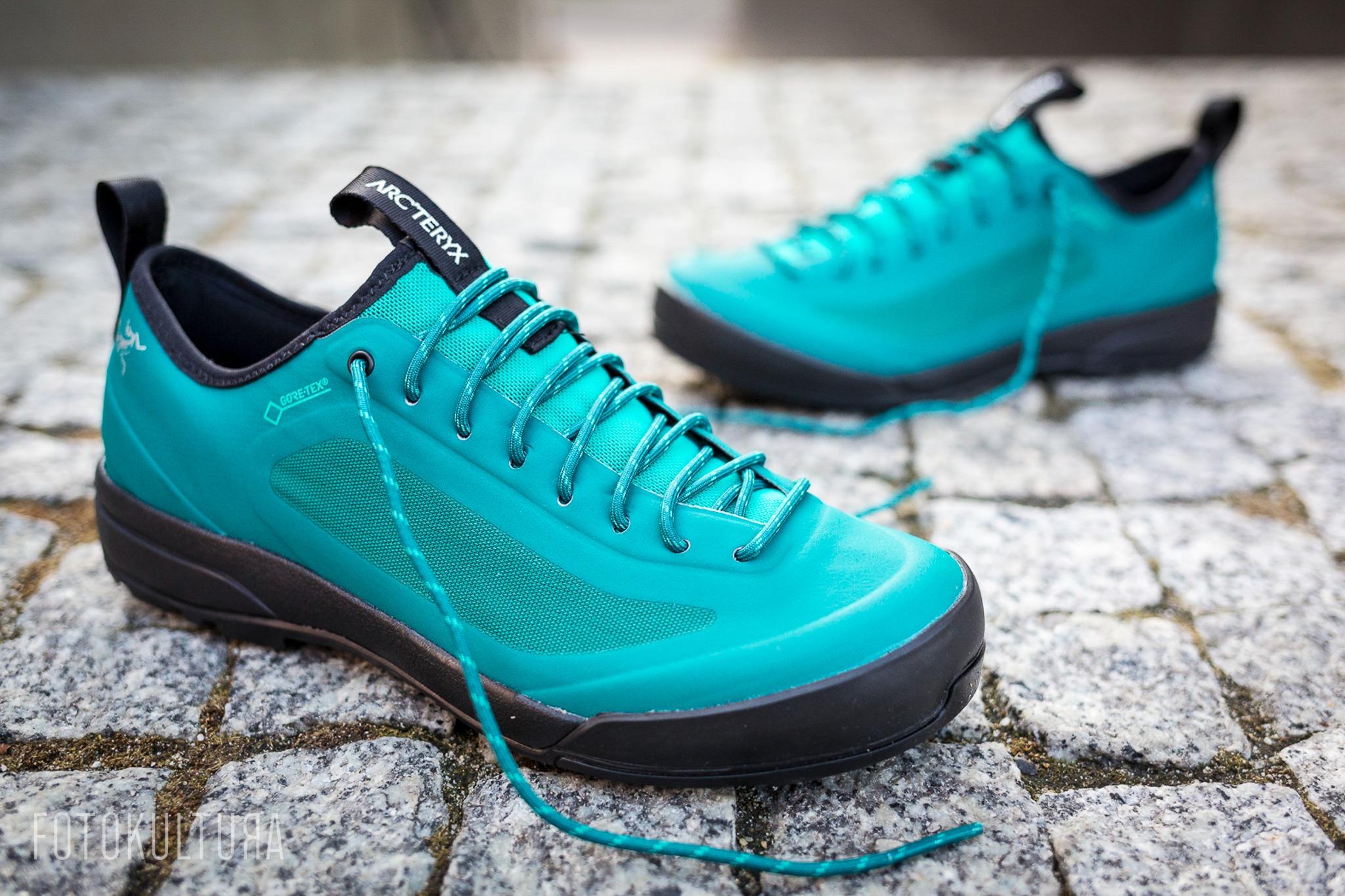 Turkusowe buty trekkingowe Arc'teryx z czarną podeszwą i sznurówkami na tle szarej kostki brukowej, widoczne logo GORE-TEX.