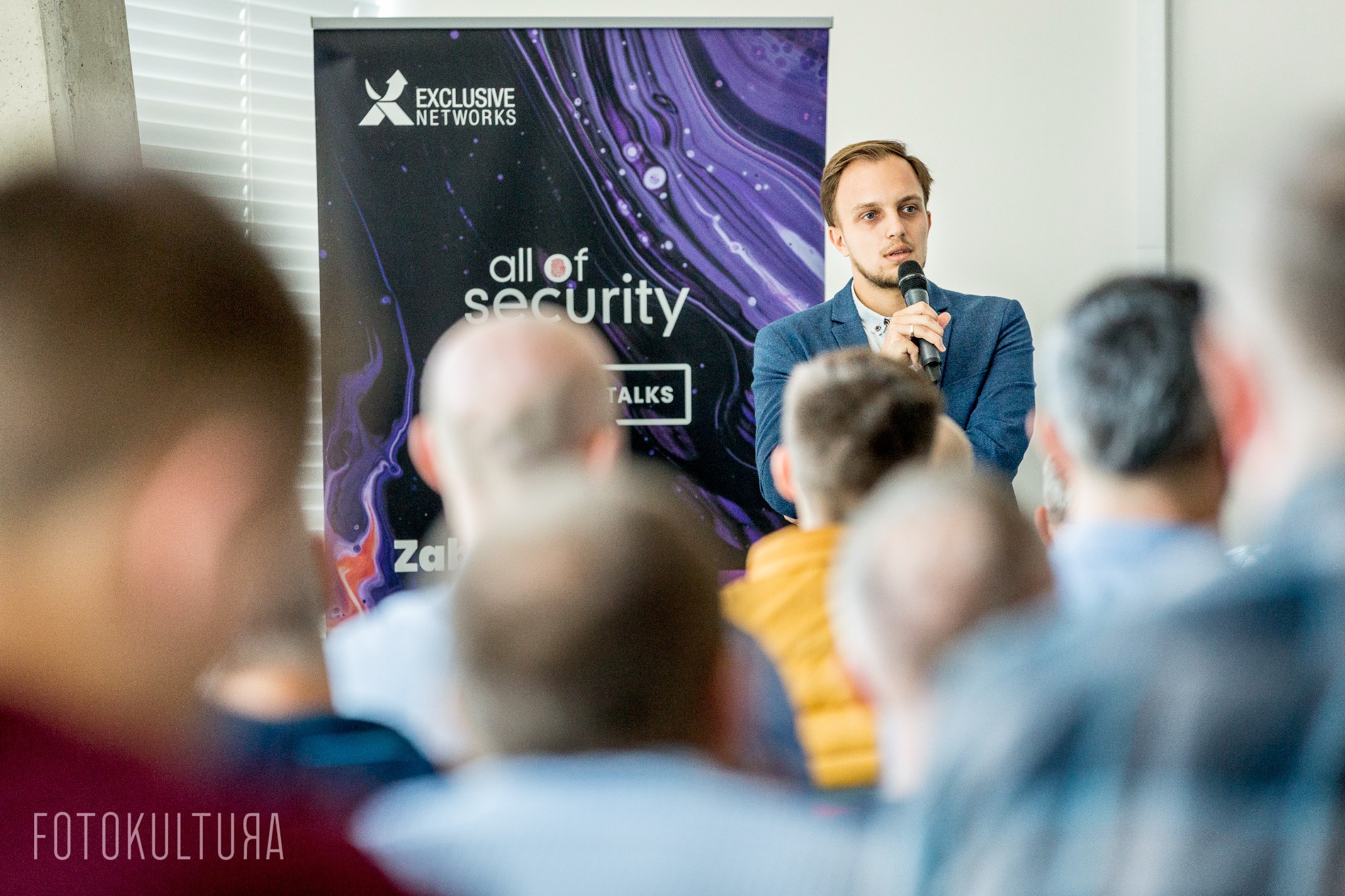 Prelegent w błękitnej marynarce przemawia do publiczności podczas eventu 'All of Security', z widocznym banerem Exclusive Networks w tle.