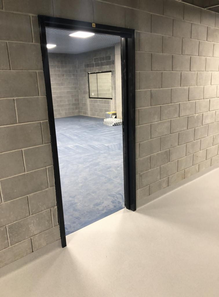 Widok przez futrynę drzwiową na pomieszczenie z niebieską posadzką, ściany z szarych bloczków betonowych, w tle okno i materiały budowlane.