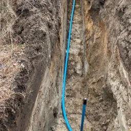 Wykop z ułożoną niebieską rurą wodociągową i pionowym elementem przyłączeniowym na dnie, widok z góry.